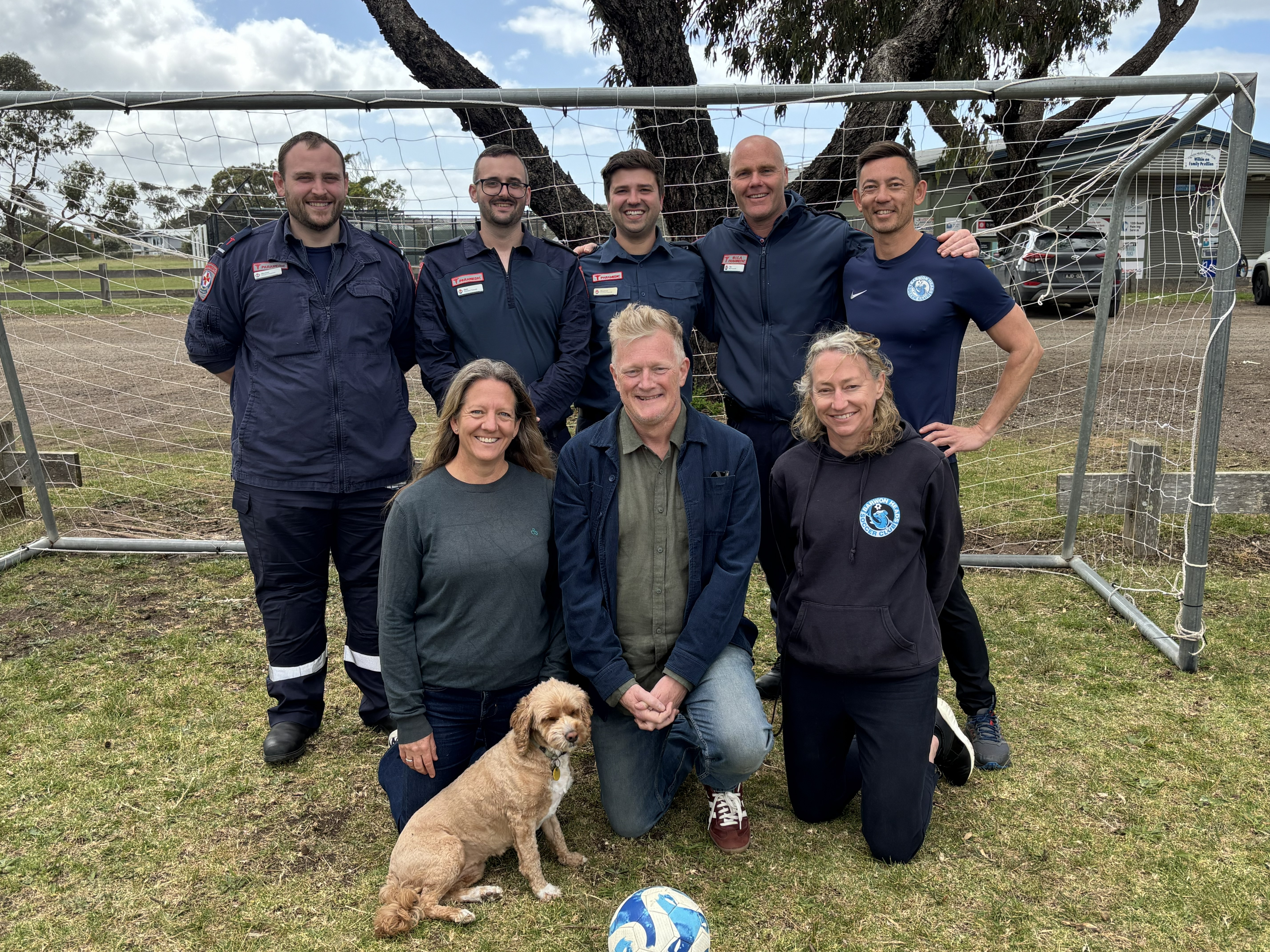 A group of four paramedics and four others in a soccer goal with a dog and soccer ball. 